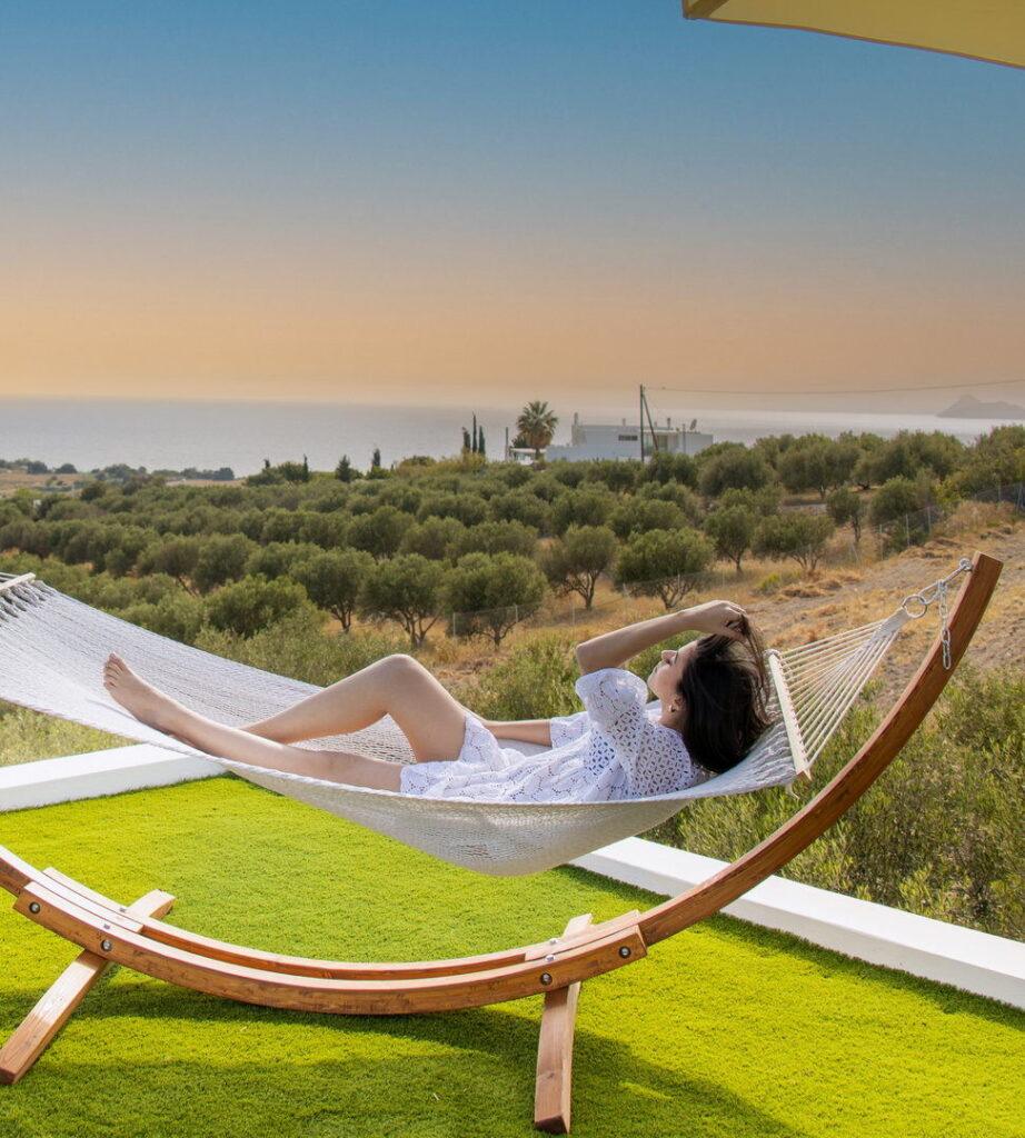 A woman relaxes in a hammock on a balcony overlooking an olive grove and the sea at sunset.
