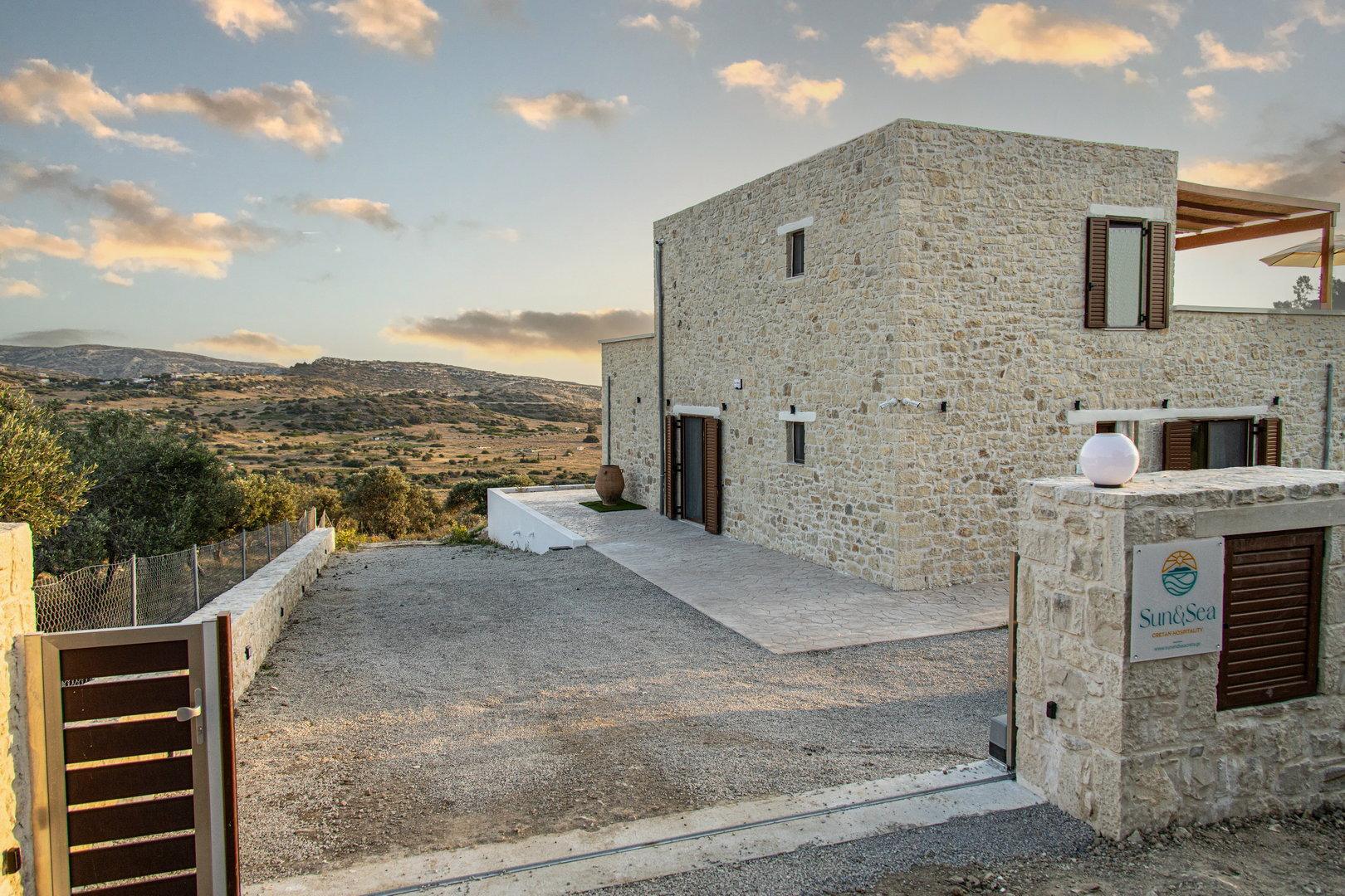 Sun and Sea villa with a gated entrance and a gravel driveway, overlooking a landscape of hills and olive trees under a cloudy sky at sunset.